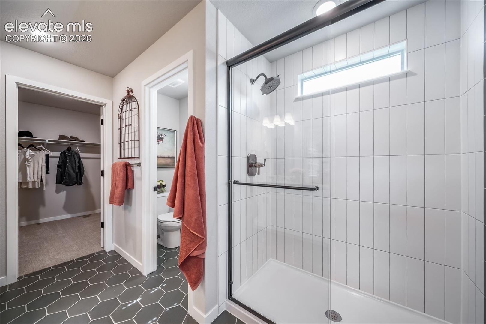 Contemporary bathroom featuring dark hexagonal floor tiles, a glass-enclosed shower with vertical white subway tile, and a transom window
*Photos of a model home*