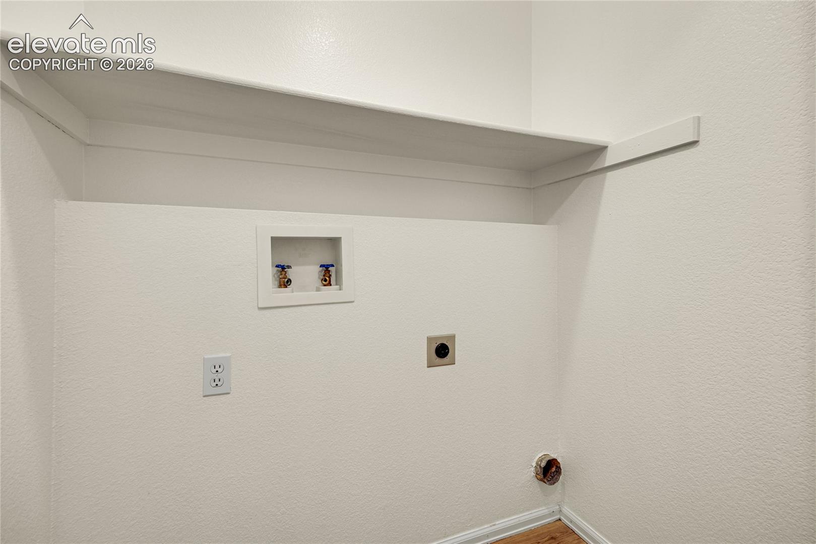 Utility area featuring a wall-mounted shelving unit, recessed plumbing connections, an integrated electrical outlet, and a dedicated exhaust vent