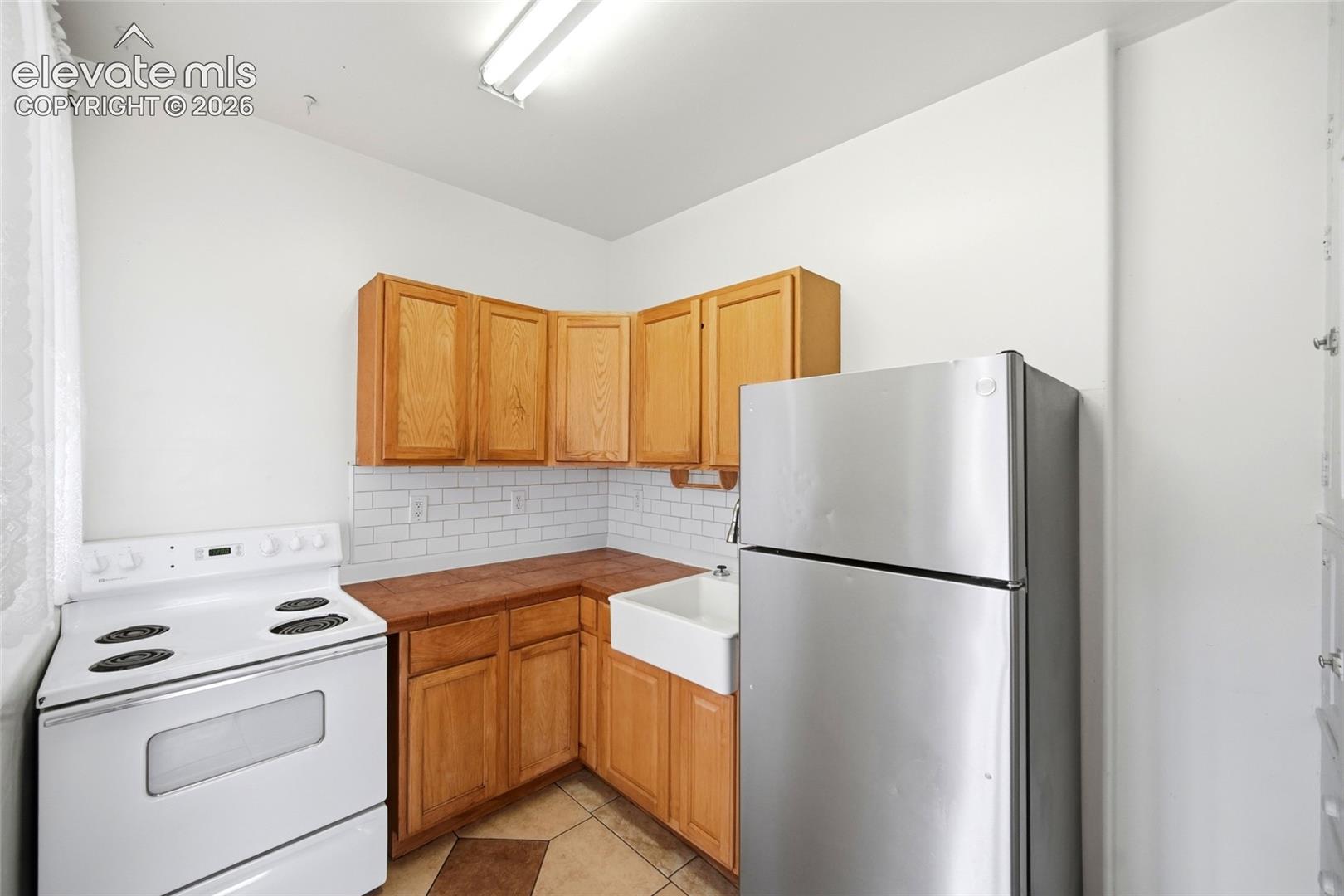Kitchen featuring natural wood cabinetry, subway tile backsplash, white farmhouse sink, and stainless steel refrigerator