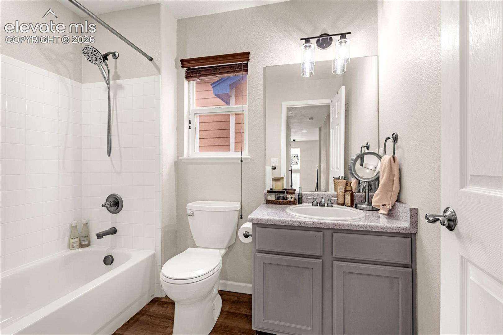 Bathroom featuring a shower-tub combination with white tile surround, vanity with stone-composite countertop, undermount sink, dark grey cabinetry, and wood-finish flooring