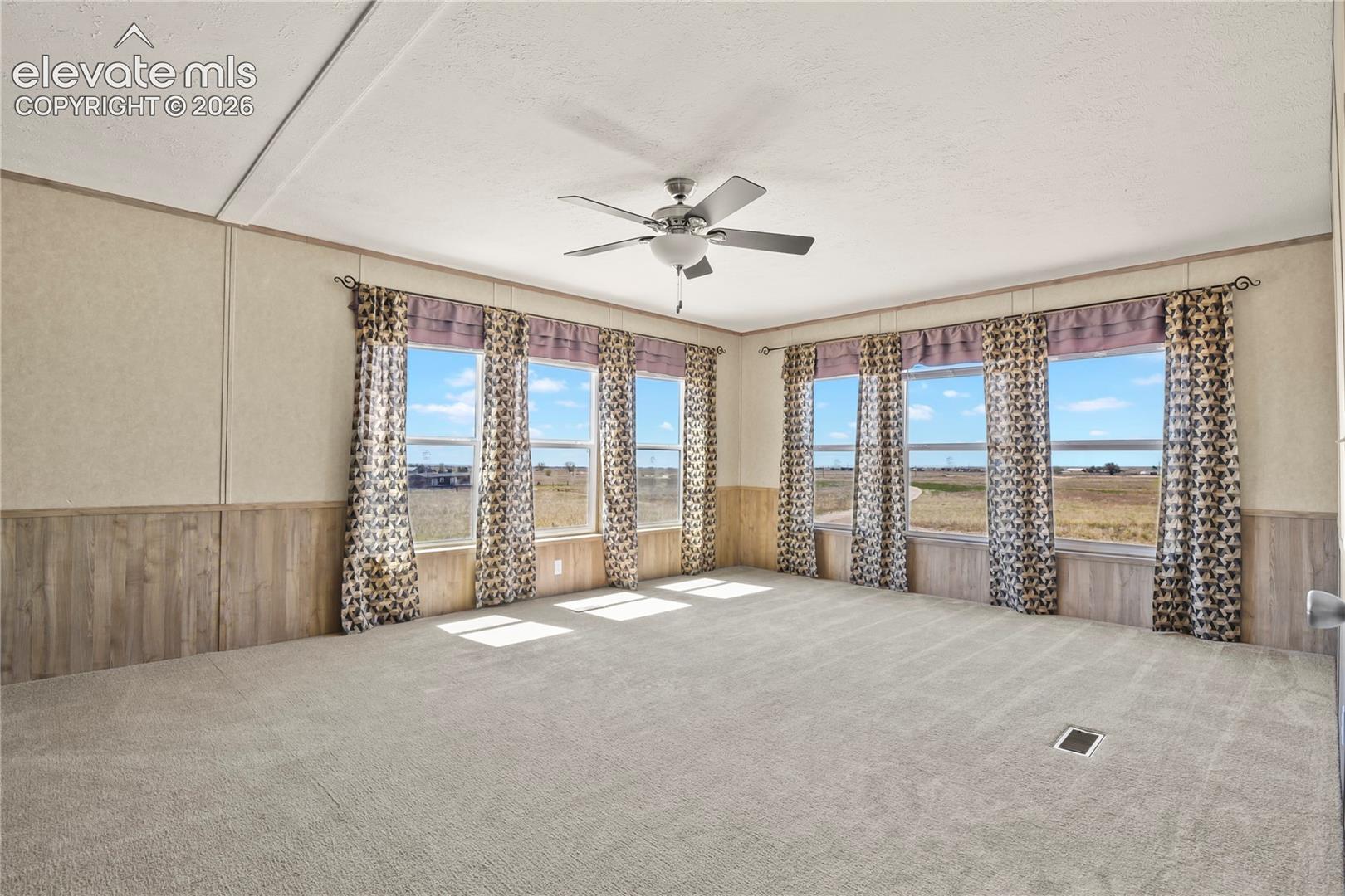 Spacious room featuring carpeting, wood-finish wainscoting, six expansive windows, and a ceiling fan with integrated lighting
