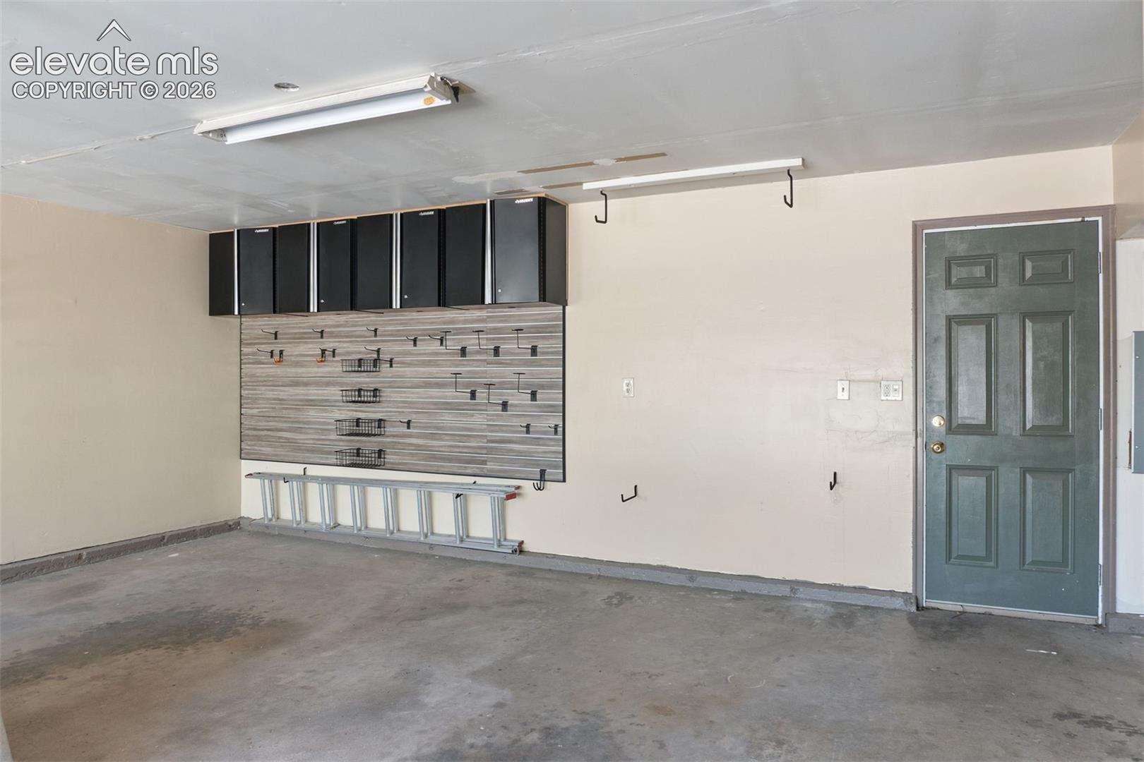 Garage space featuring wall-mounted black cabinetry, an integrated slatwall storage system, ceiling-mounted fluorescent lighting, and a solid six-panel entry door