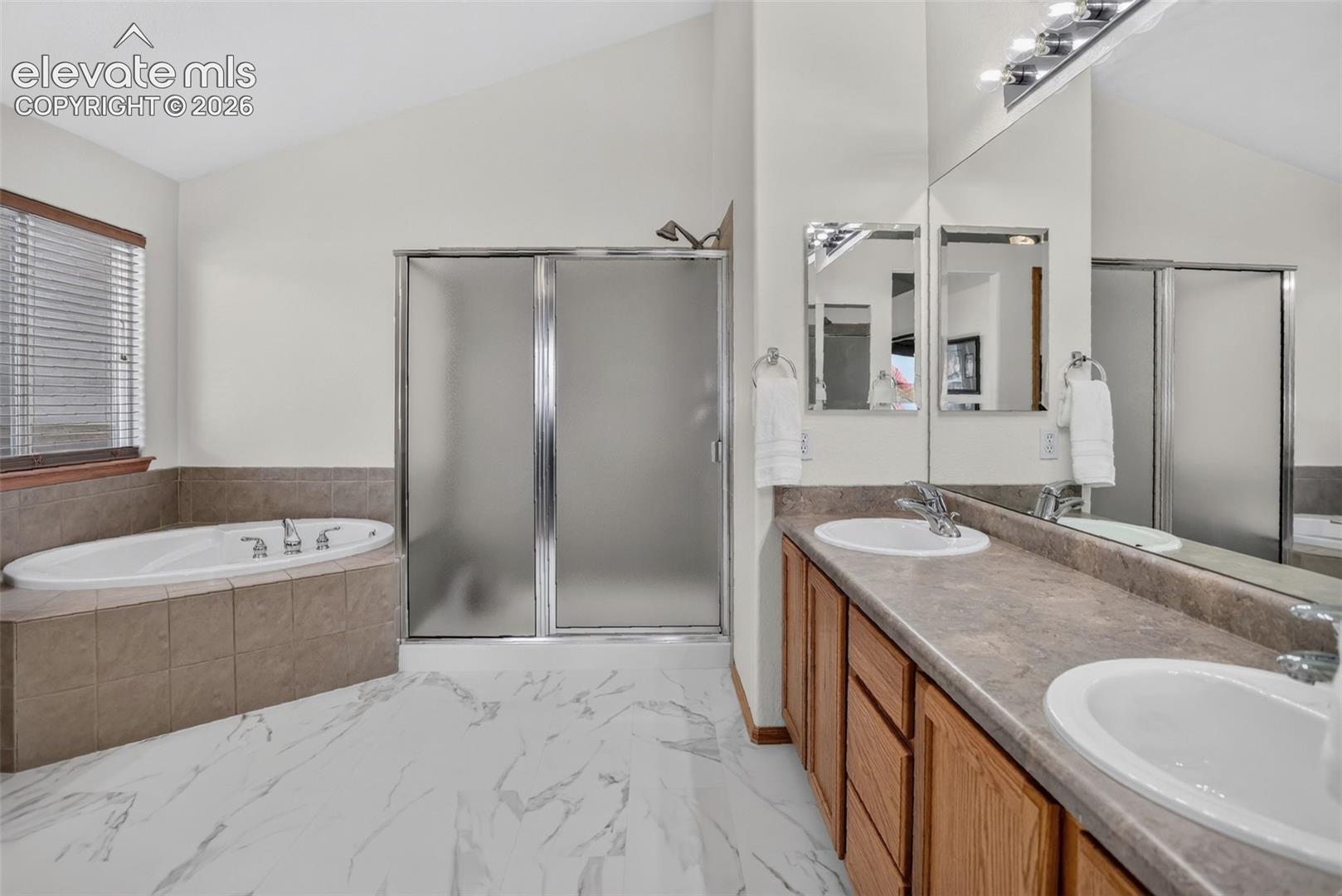 Bathroom featuring a vaulted ceiling, dual vanity with undermount sinks, a large mirror, a tiled soaking tub, and a glass-enclosed shower