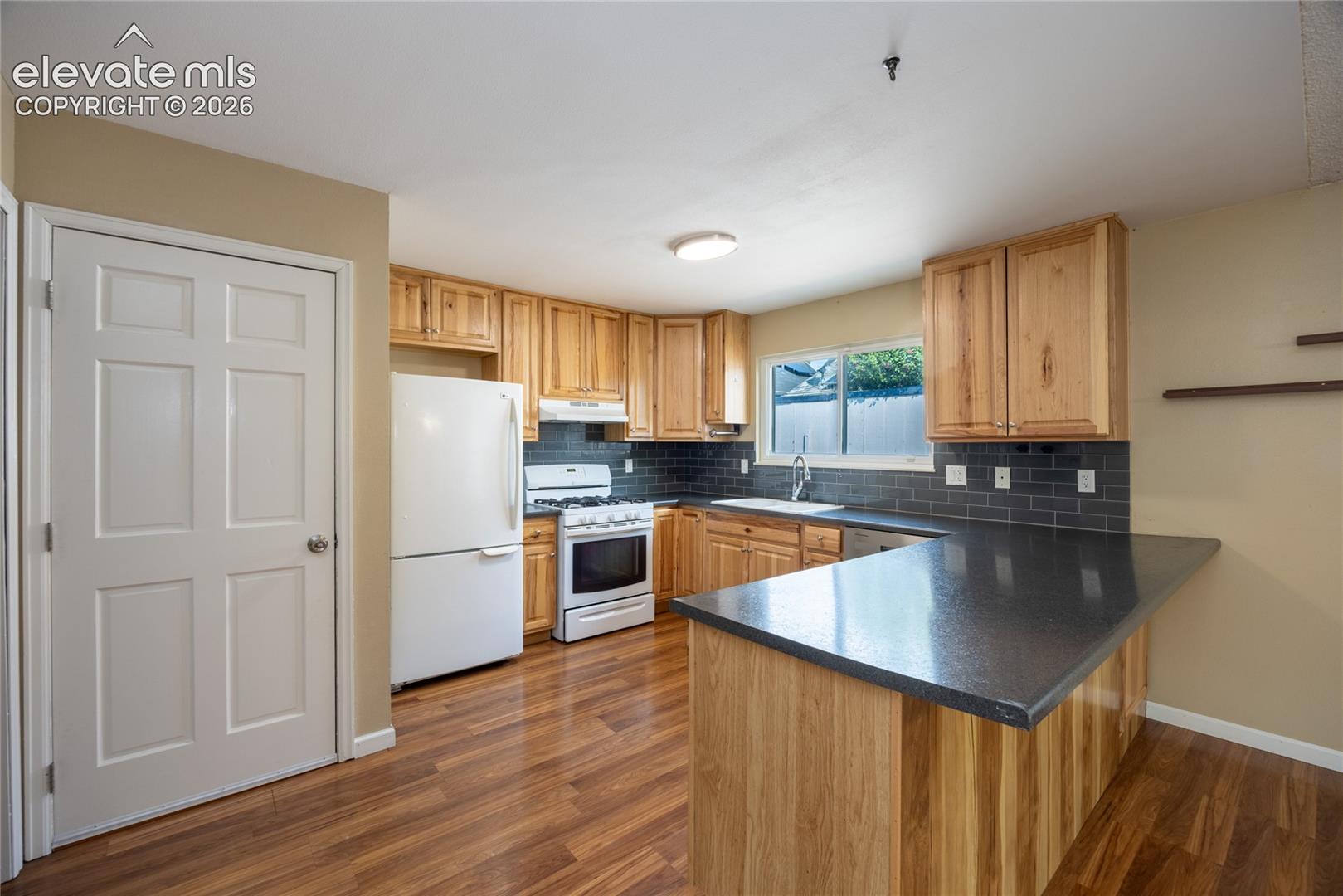 Kitchen featuring wood-finish flooring, natural wood cabinetry, dark subway tile backsplash, and dark countertops