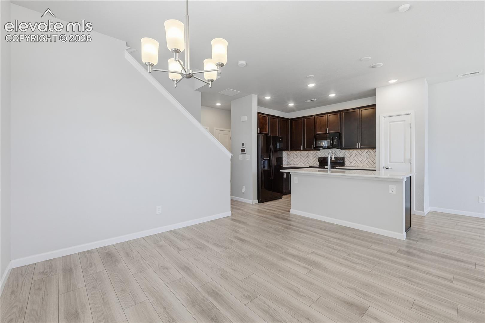 Open-concept living space featuring wood-finish flooring, a modern chandelier, and a kitchen with dark cabinetry, black appliances, and a herringbone tile backsplash