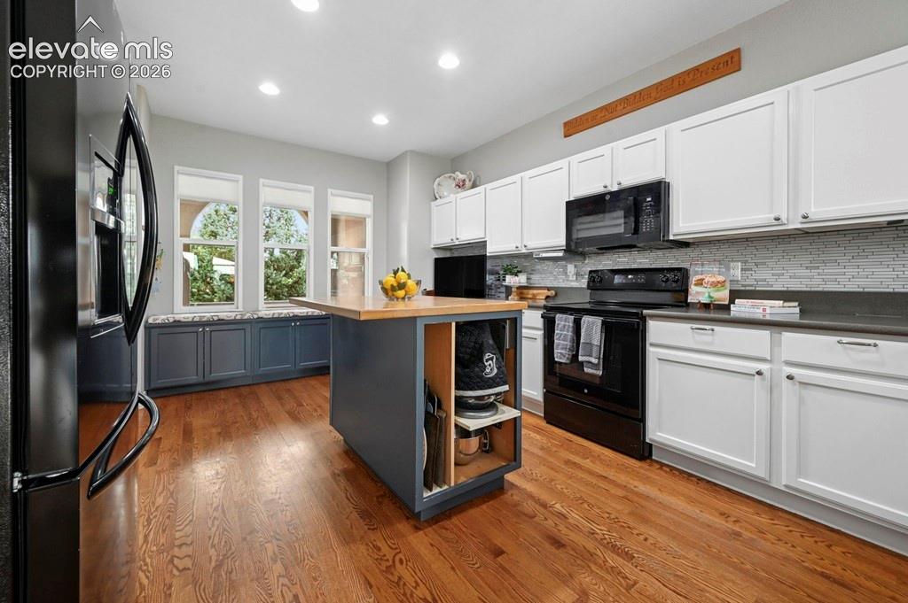 Hardwood flooring, kitchen island with wood-finish countertop, white upper and lower cabinetry, dark countertop surfaces, tiled backsplash