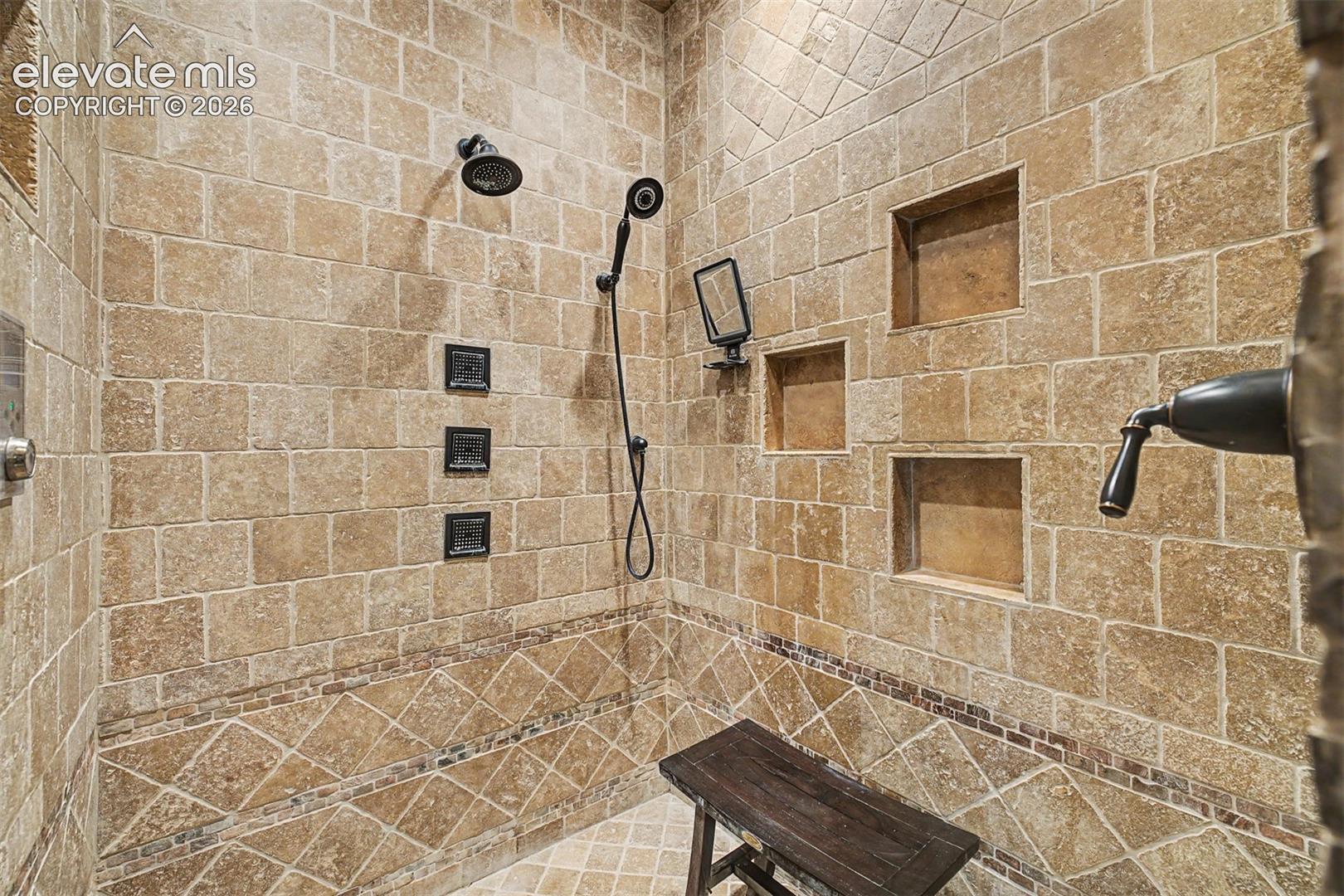 The primary suite's walk-in shower is fully wrapped in travertine tile, with a built-in body spray system, multiple storage niches, and a seated bench — a true spa-style retreat within the home.