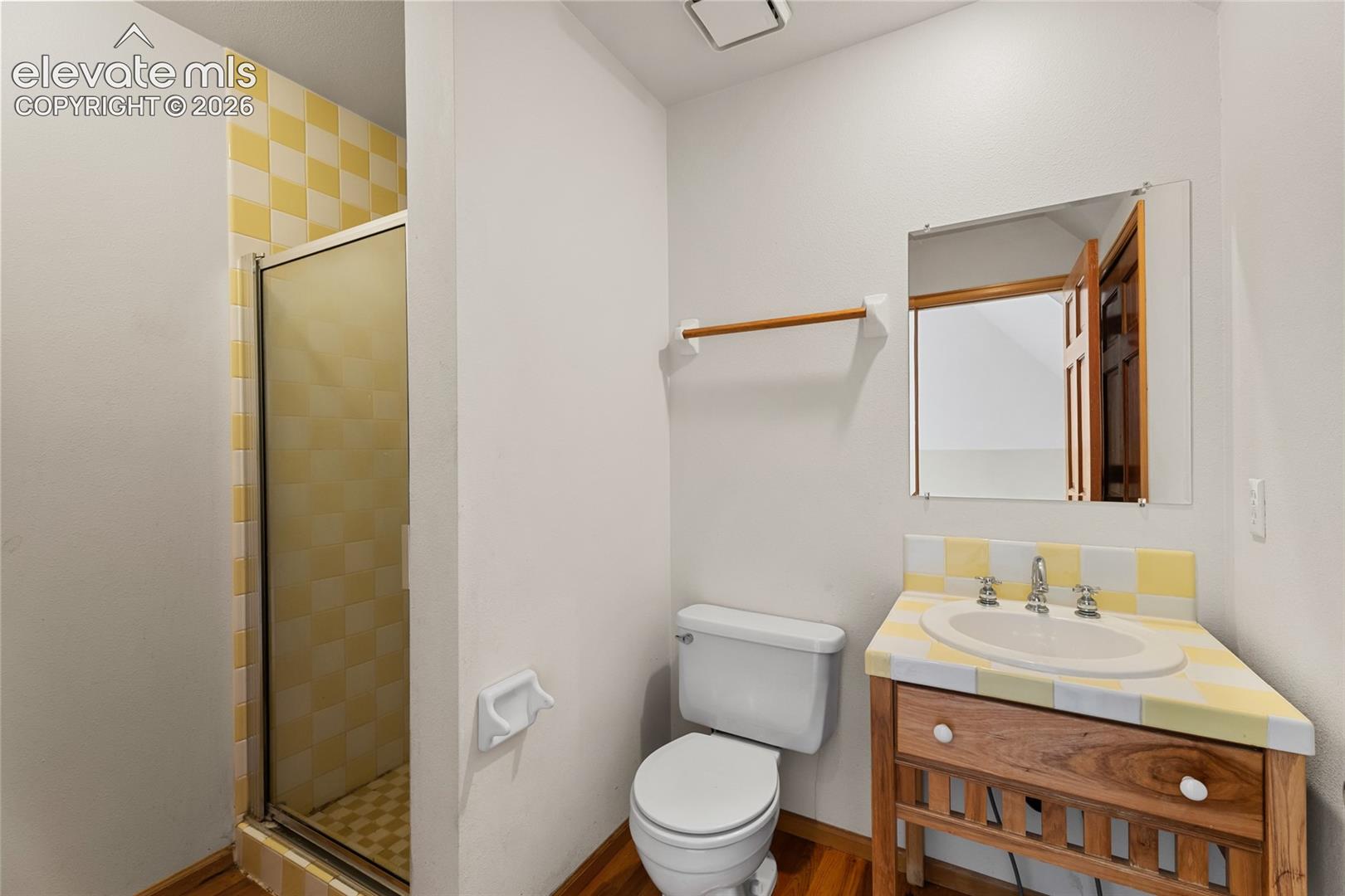 Shower enclosure with checkered yellow and white tile, a built-in vanity with a white basin sink, a wall-mounted mirror, and wood-finish flooring