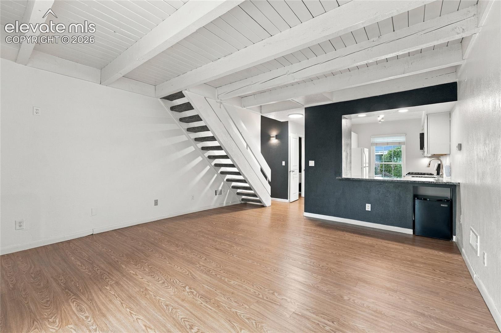 Interior featuring exposed white-painted wood-beam ceilings, wood-finish flooring, a built-in open staircase with white railing, and a pass-through kitchen counter with a stone-surface finish