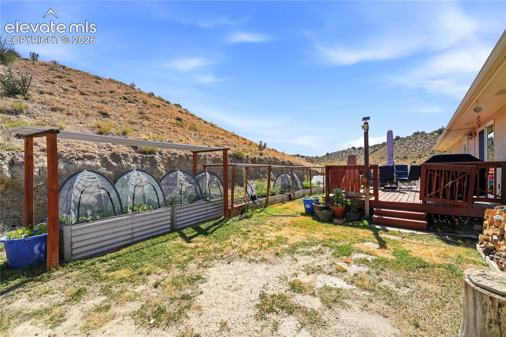 Expansive outdoor space featuring a wood deck, hillside backdrop, raised garden beds with covers, and a wood privacy fence