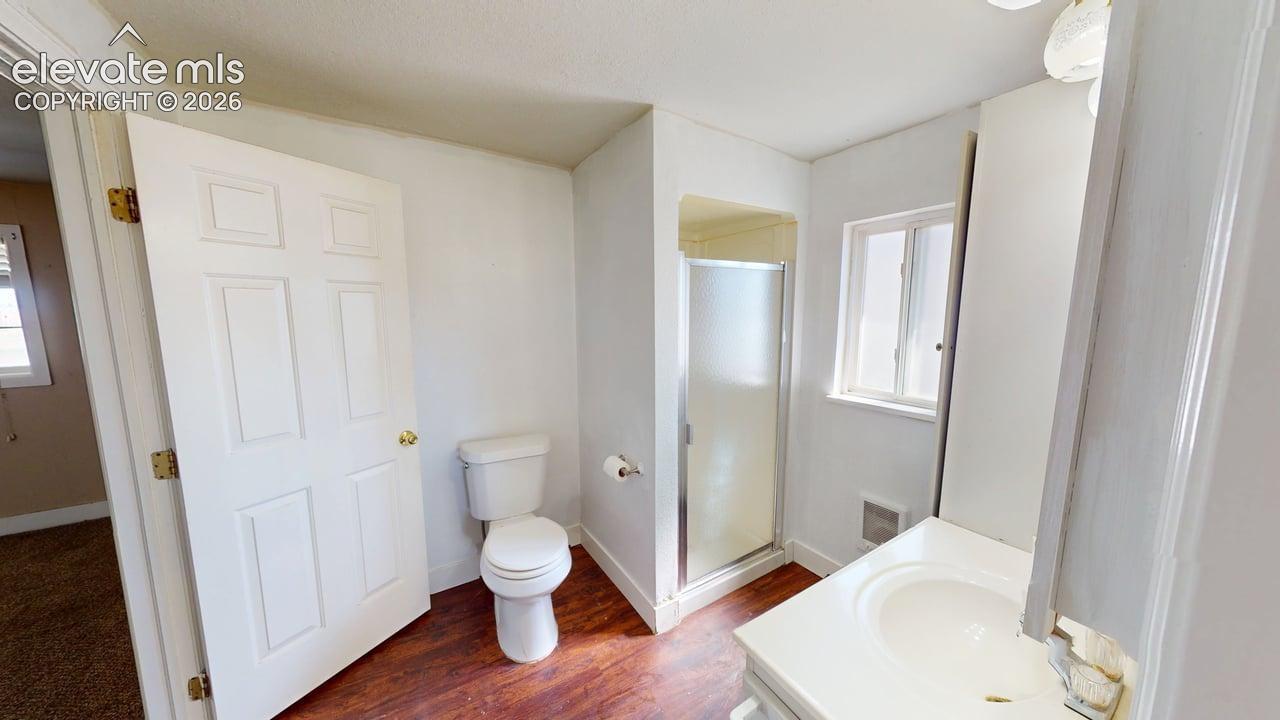 White paneled door, wood-finish flooring, white toilet, glass-enclosed shower, and a single window