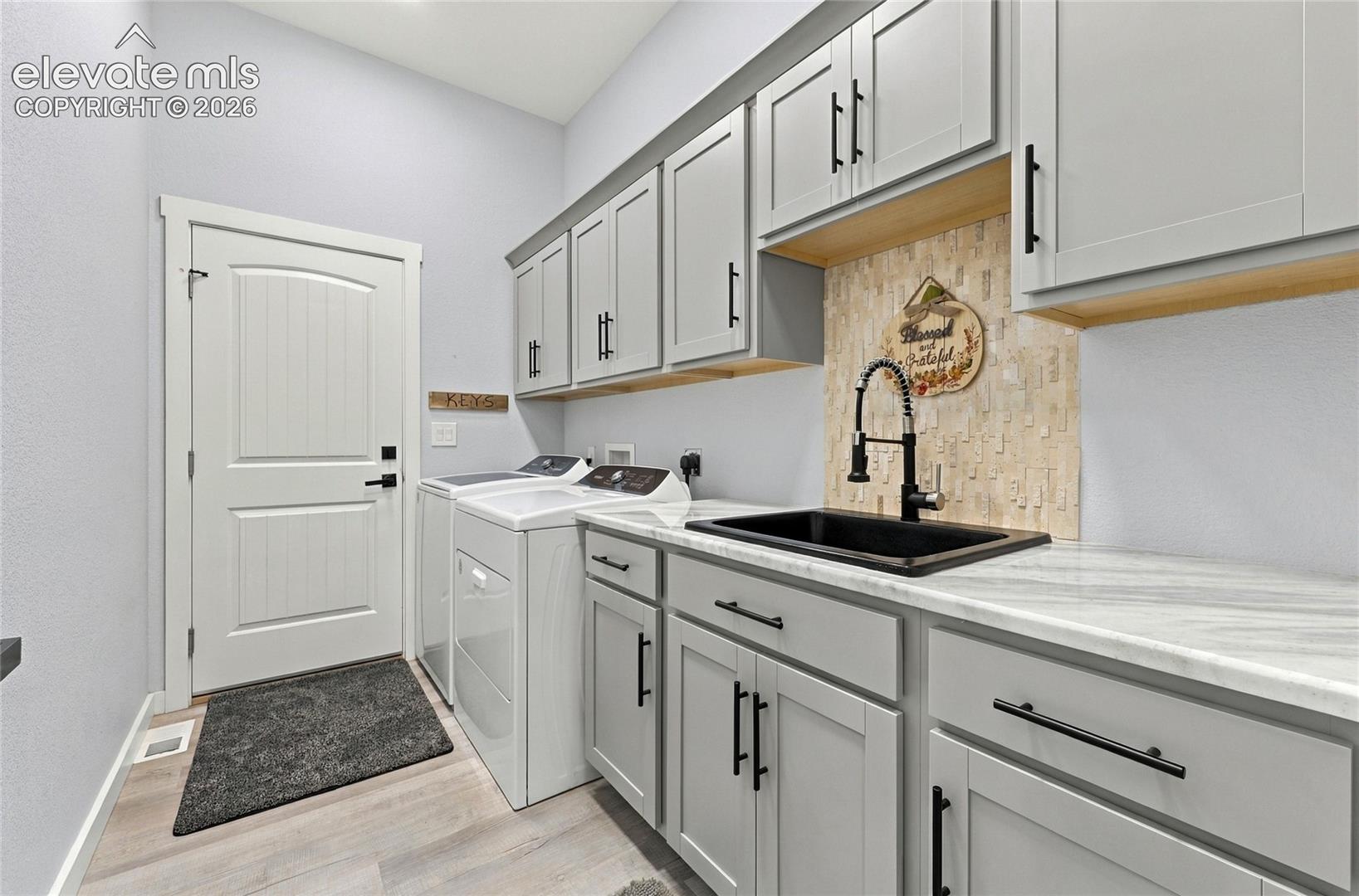 Laundry room featuring shaker-style grey cabinetry, neutral stone-patterned countertops, a drop-in black basin sink with a high-arch matte black faucet, and a tiled backsplash