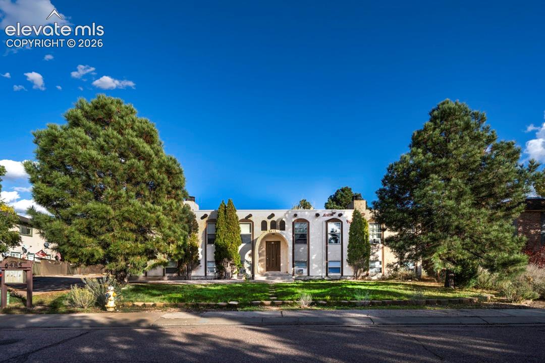 Stucco exterior featuring arched entryways, decorative protruding vigas, multi-story vertical window banks, and mature pine landscaping