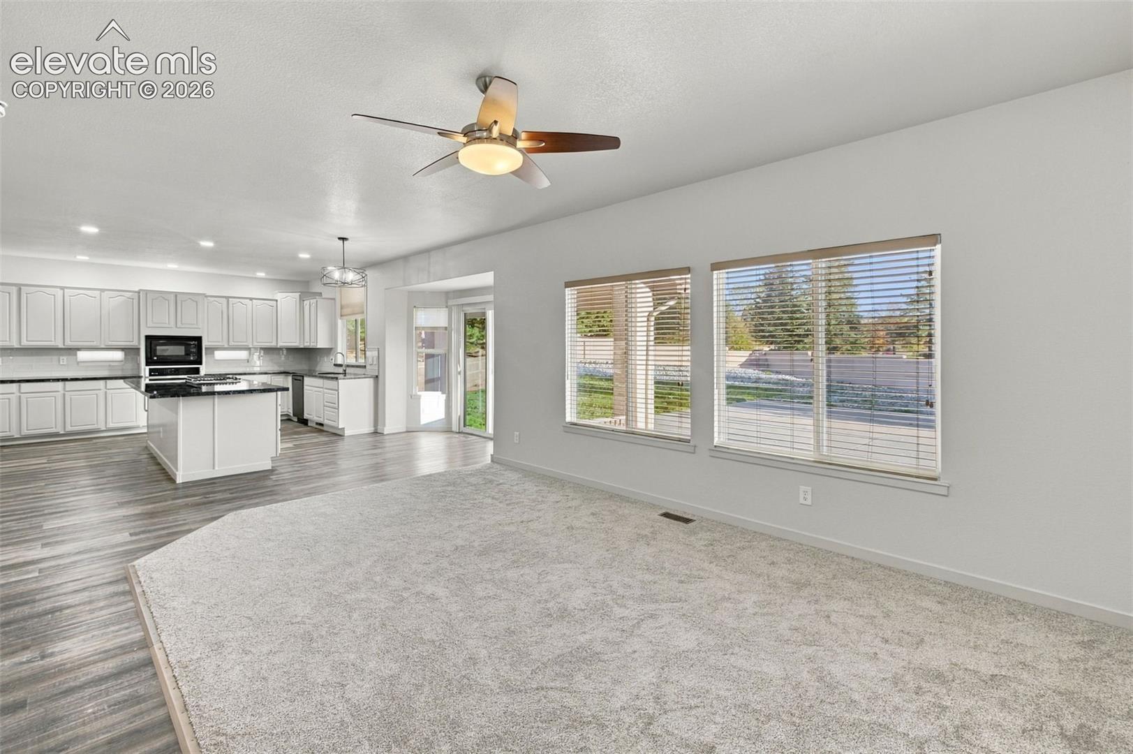 Open-concept floor plan featuring wood-finish flooring, central kitchen island with dark countertops, white cabinetry, recessed lighting, and large windows with integrated blinds