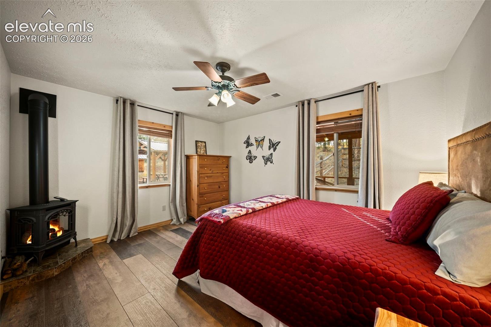 Wood-finish flooring, freestanding black metal stove, stone hearth, ceiling fan with integrated lighting, and dual windows