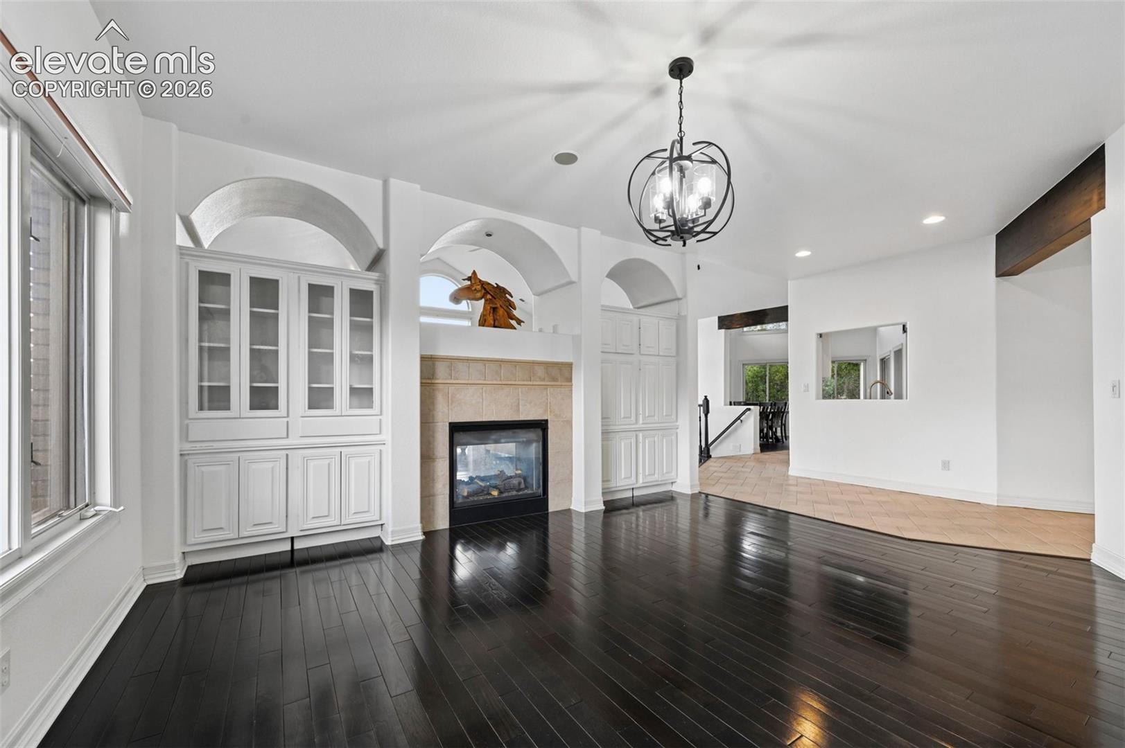 Dark wood-finish flooring, built-in cabinetry with glass display doors, arched architectural details, a stone-clad fireplace, and a hanging chandelier