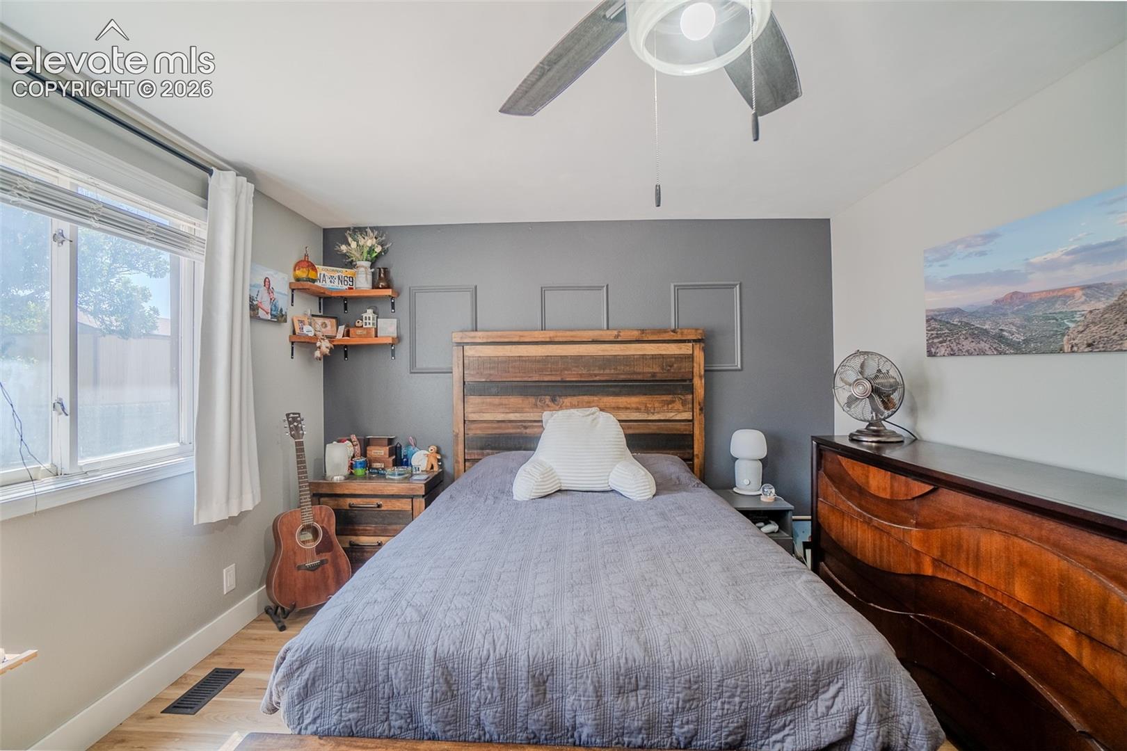 Bedroom featuring wood-finish flooring, a window with blinds, a ceiling fan with light fixture, and a feature wall with a contrasting paint color