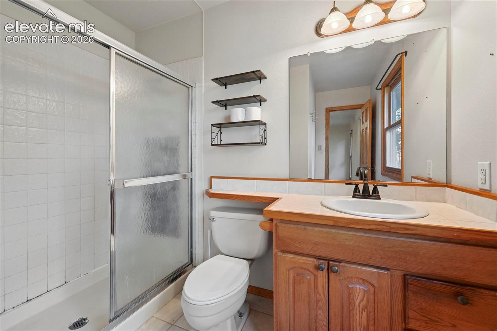 Wood-finish vanity with an integrated sink, a three-light vanity fixture, a sliding glass shower enclosure, and white tiled walls