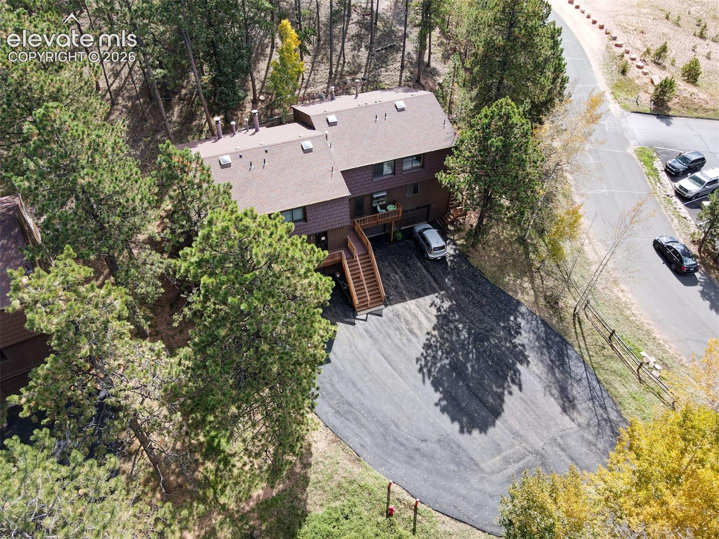 Multi-level brown-sided residence with multiple rooflines, a dark asphalt driveway, a visible parking pad, and mature surrounding trees