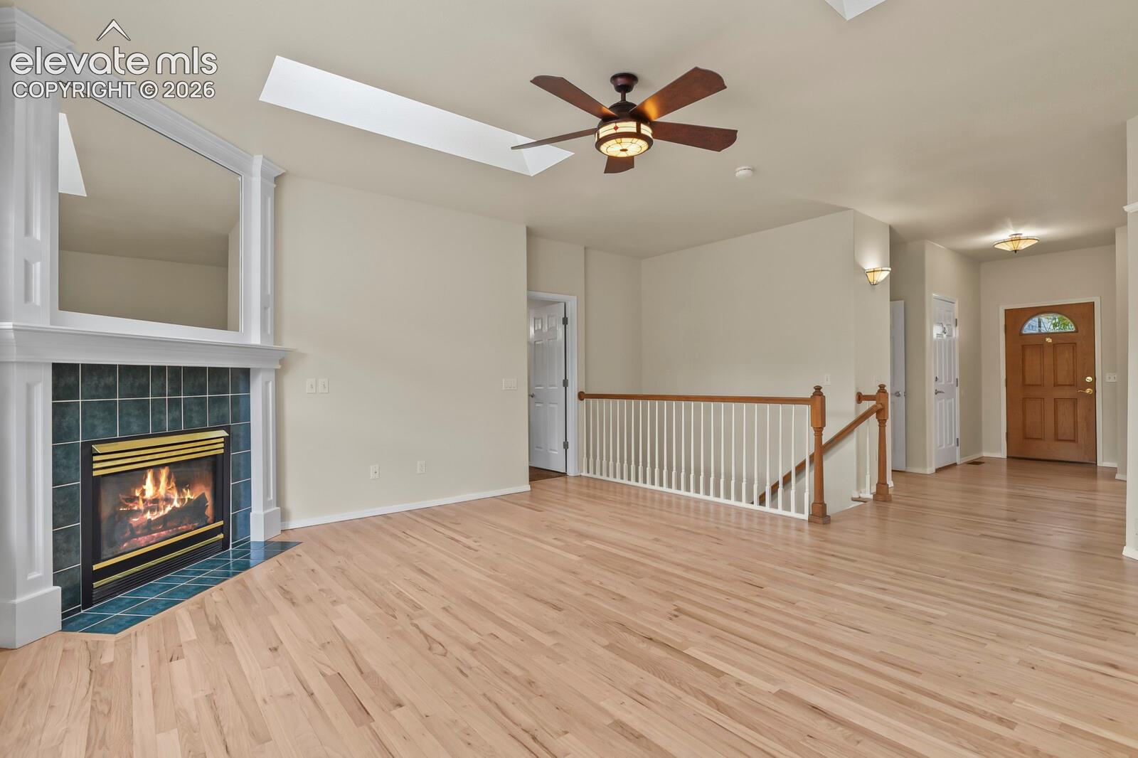 Fireplace featuring a tiled surround and white mantel, wood-finish flooring, skylight, ceiling fan with integrated lighting, and a wooden stair railing