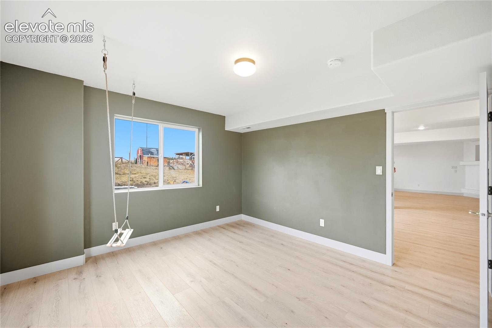 Neutral-toned interior room featuring wood-finish flooring, a large window, flush-mount ceiling lighting, and a ceiling-mounted swing