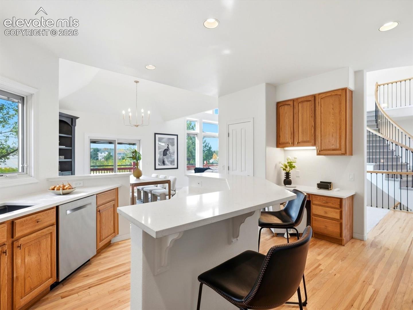 Kitchen featuring a peninsula with solid-surface countertops, light wood cabinetry, stainless steel dishwasher, wood-finish flooring, and a curved staircase with a metal railing