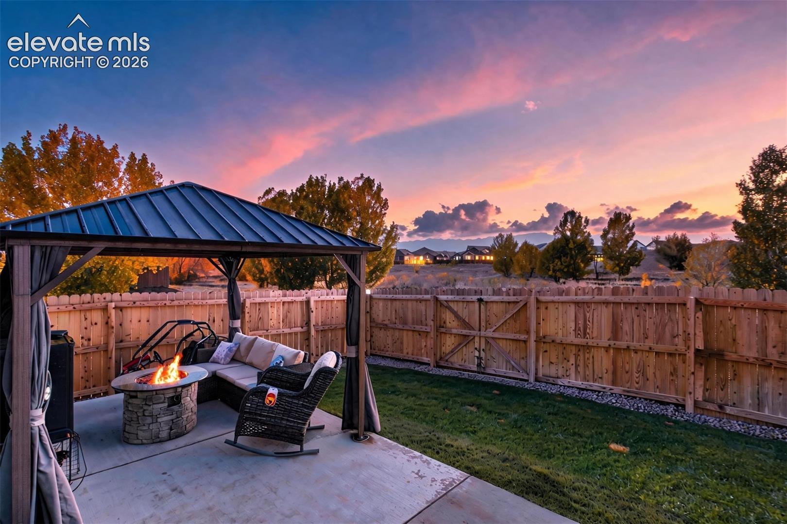 Covered metal-roofed gazebo on a concrete patio, featuring a stone-clad fire pit and a wood privacy fence