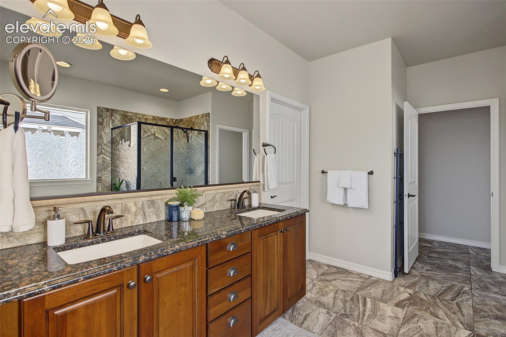 Dual-sink vanity with stone-finish countertops, warm-toned wood cabinetry, large vanity mirror with multi-light fixtures, and tiled flooring