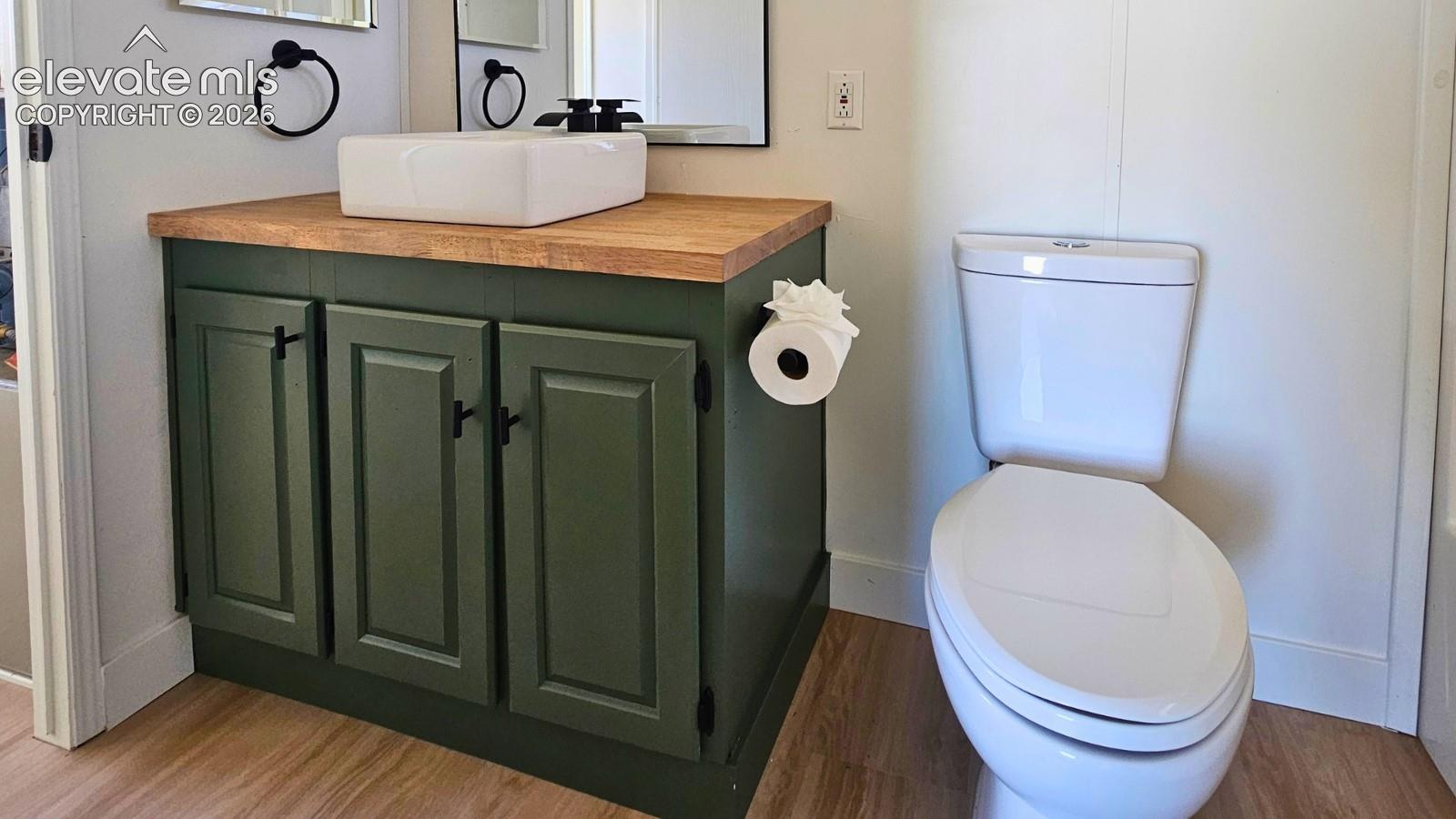 Olive-colored cabinetry with a wood-finish countertop, square vessel sink, matte black hardware, and wood-finish flooring