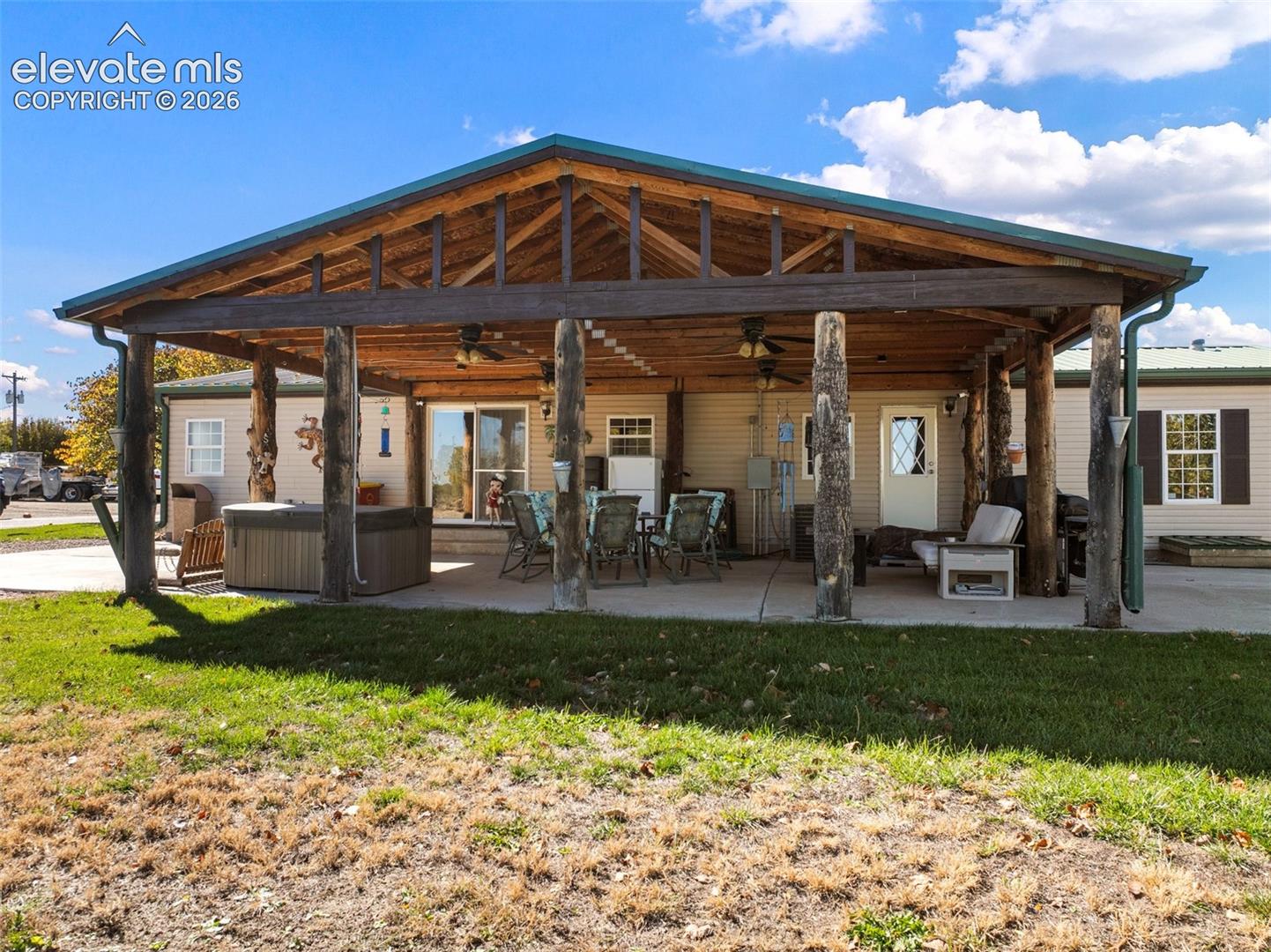 Covered outdoor patio featuring rustic log support columns, exposed timber rafters, a concrete floor, built-in ceiling fans, and an integrated spa