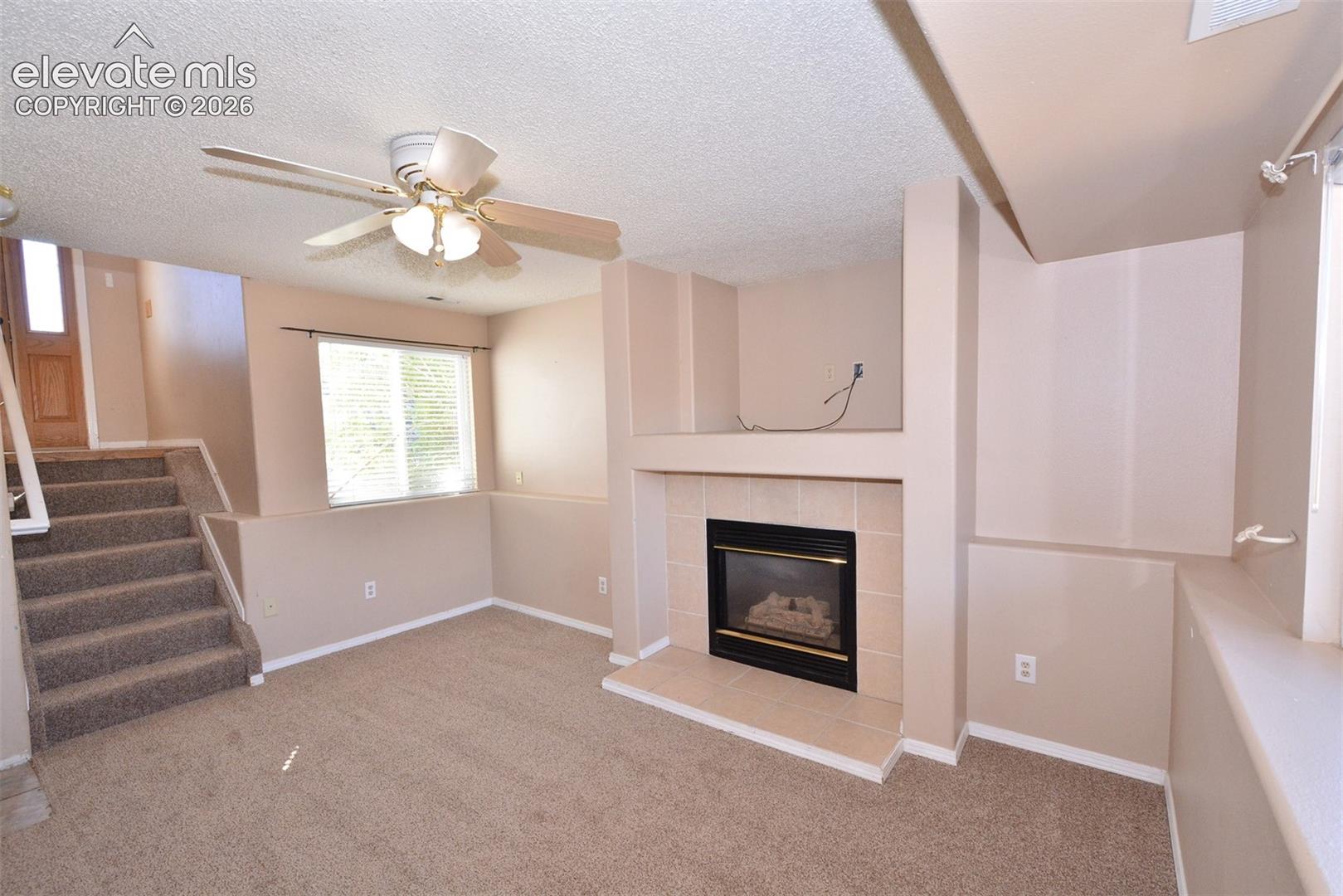 Carpeted living space featuring a built-in fireplace with tile surround, a window with blinds, a ceiling fan, and carpeted stairs with a white railing