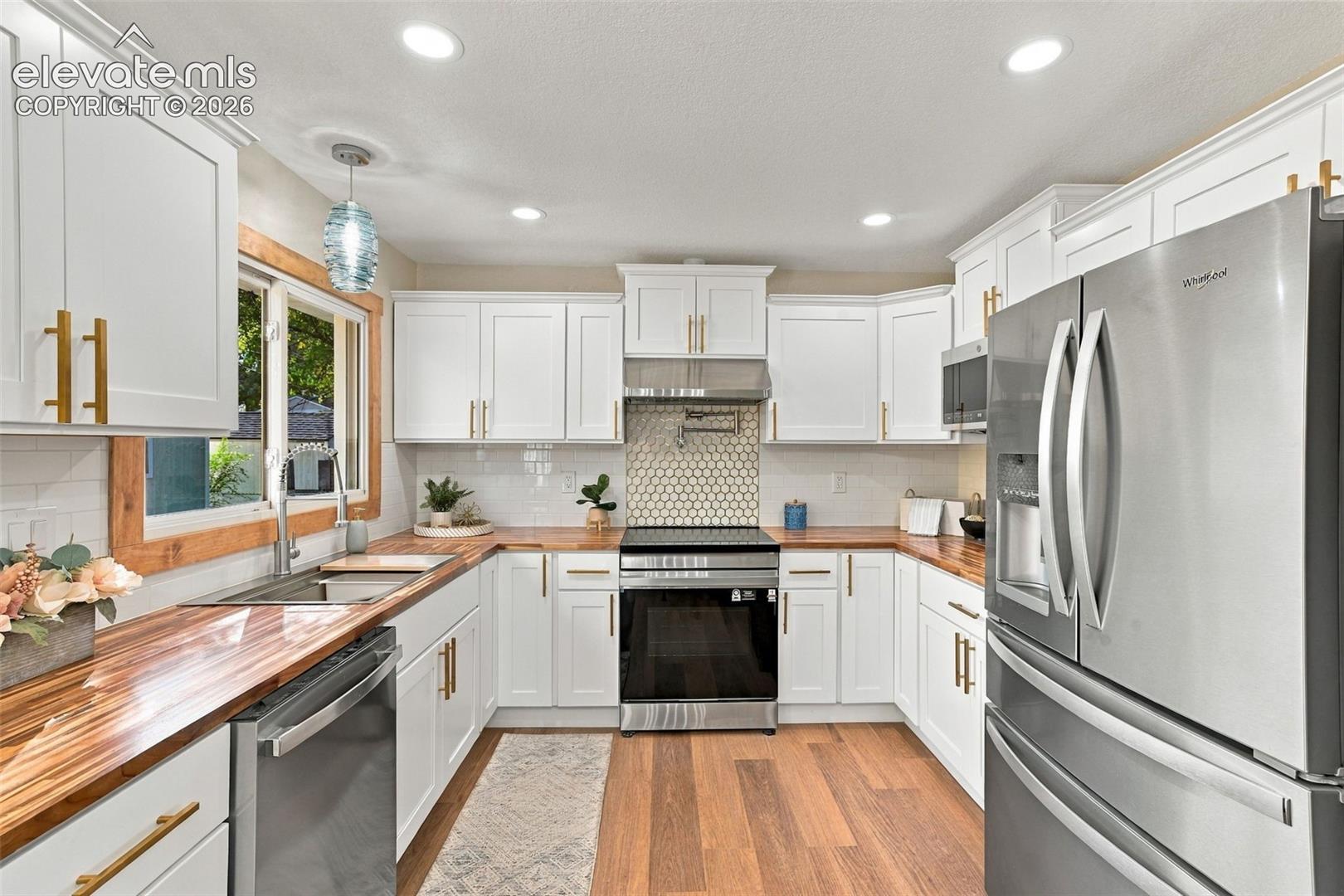 Kitchen featuring white shaker-style cabinetry with gold-tone hardware, wood-finish countertops, stainless steel appliances, a honeycomb-pattern tile backsplash, and wood-finish flooring
