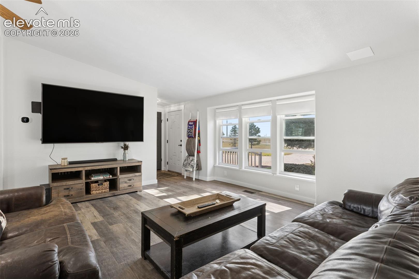 Living space featuring wood-finish flooring, a wall-mounted ceiling fan, and large windows with horizontal blinds