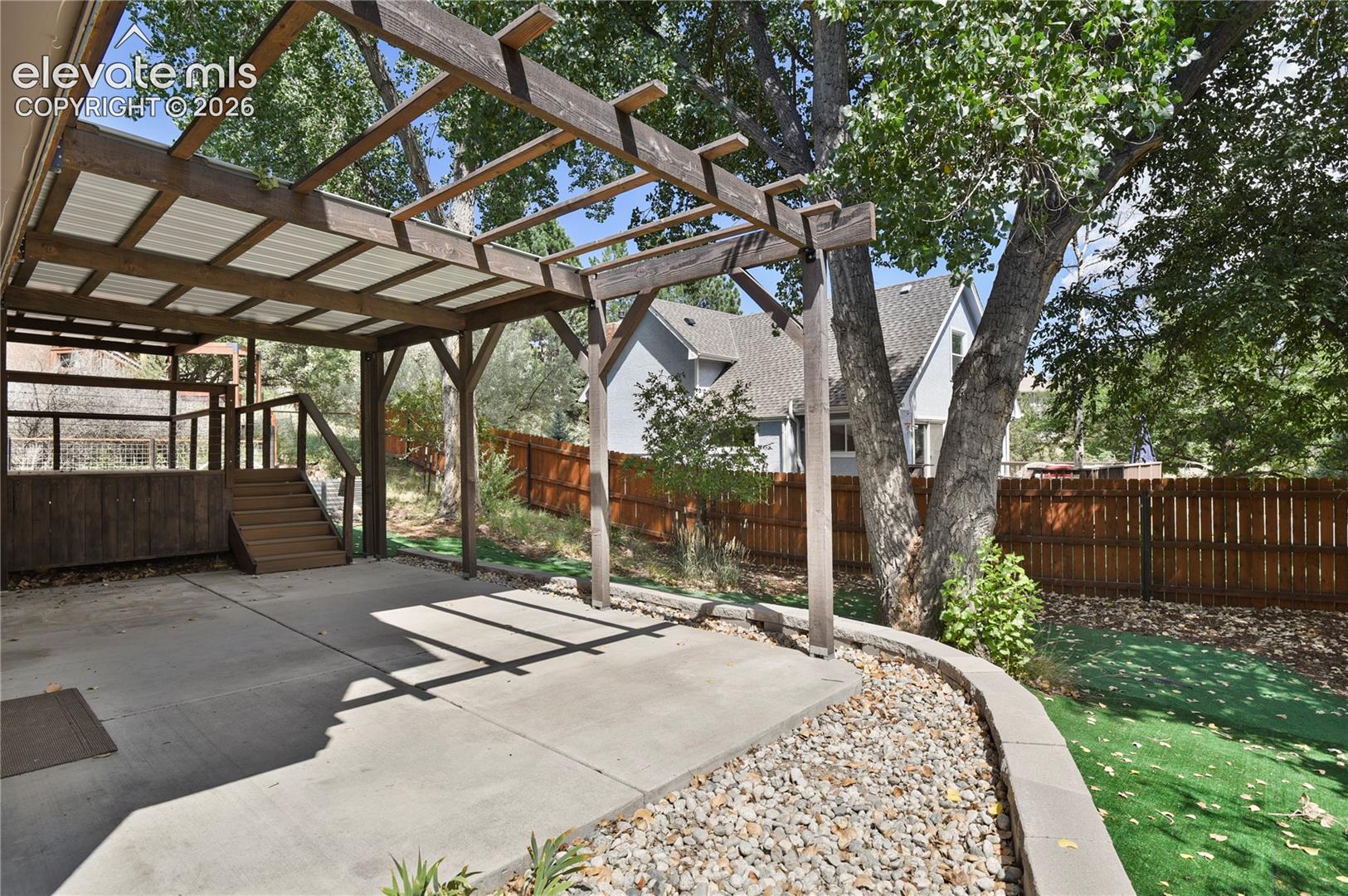 Covered wood pergola, concrete patio, elevated wooden deck with stairs, natural stone retaining wall, and fenced perimeter