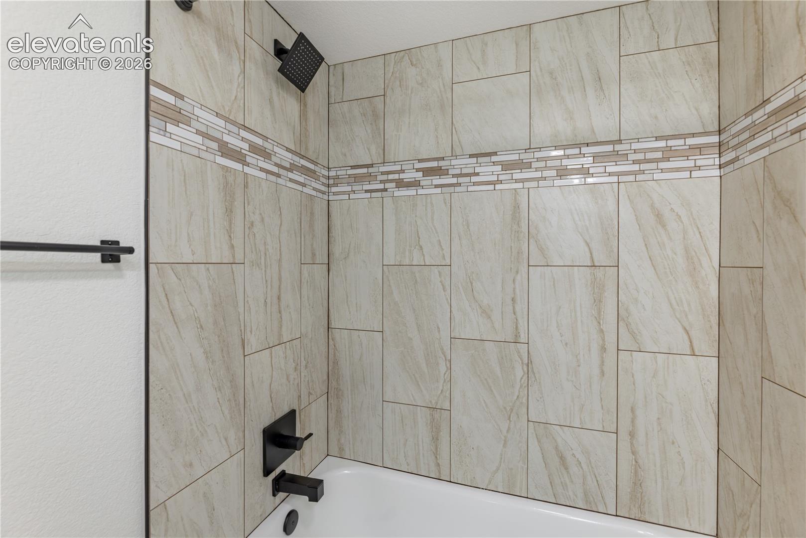 Bathtub surround featuring large-format neutral-tone wall tile, horizontal mosaic accent banding, matte black showerhead, matching matte black faucet controls, and a wall-mounted towel bar