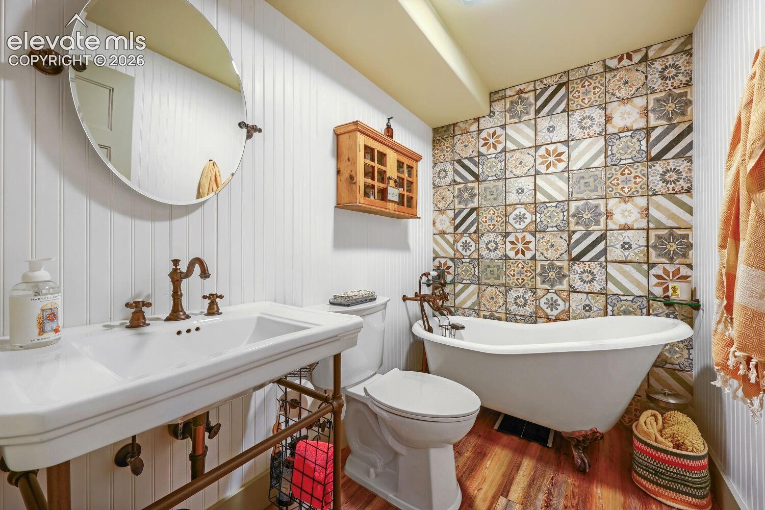 White beadboard paneling, patterned accent wall tile, clawfoot bathtub, pedestal sink with integrated basin, and wood-finish flooring