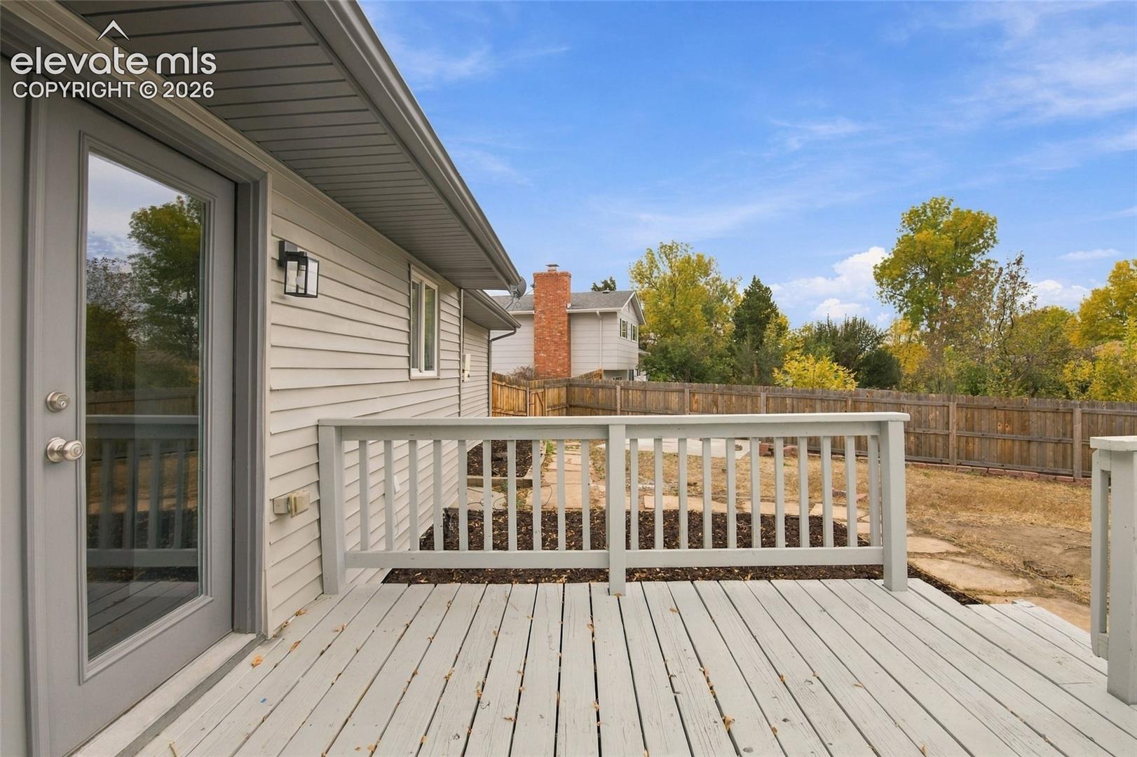 Raised exterior deck featuring painted wood-finish decking, matching balustrade railings, horizontal lap siding, and a glass-panel door with a wall-mounted exterior light fixture