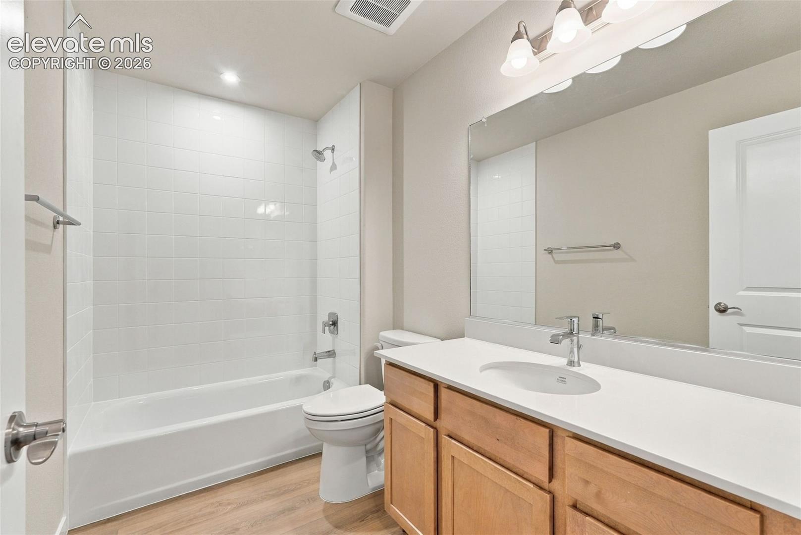 Bathroom featuring a tub-shower combination with white tile surround, light wood-finish vanity cabinet, solid surface countertop with integrated oval sink, and a large-format vanity mirror with overhead light fixture