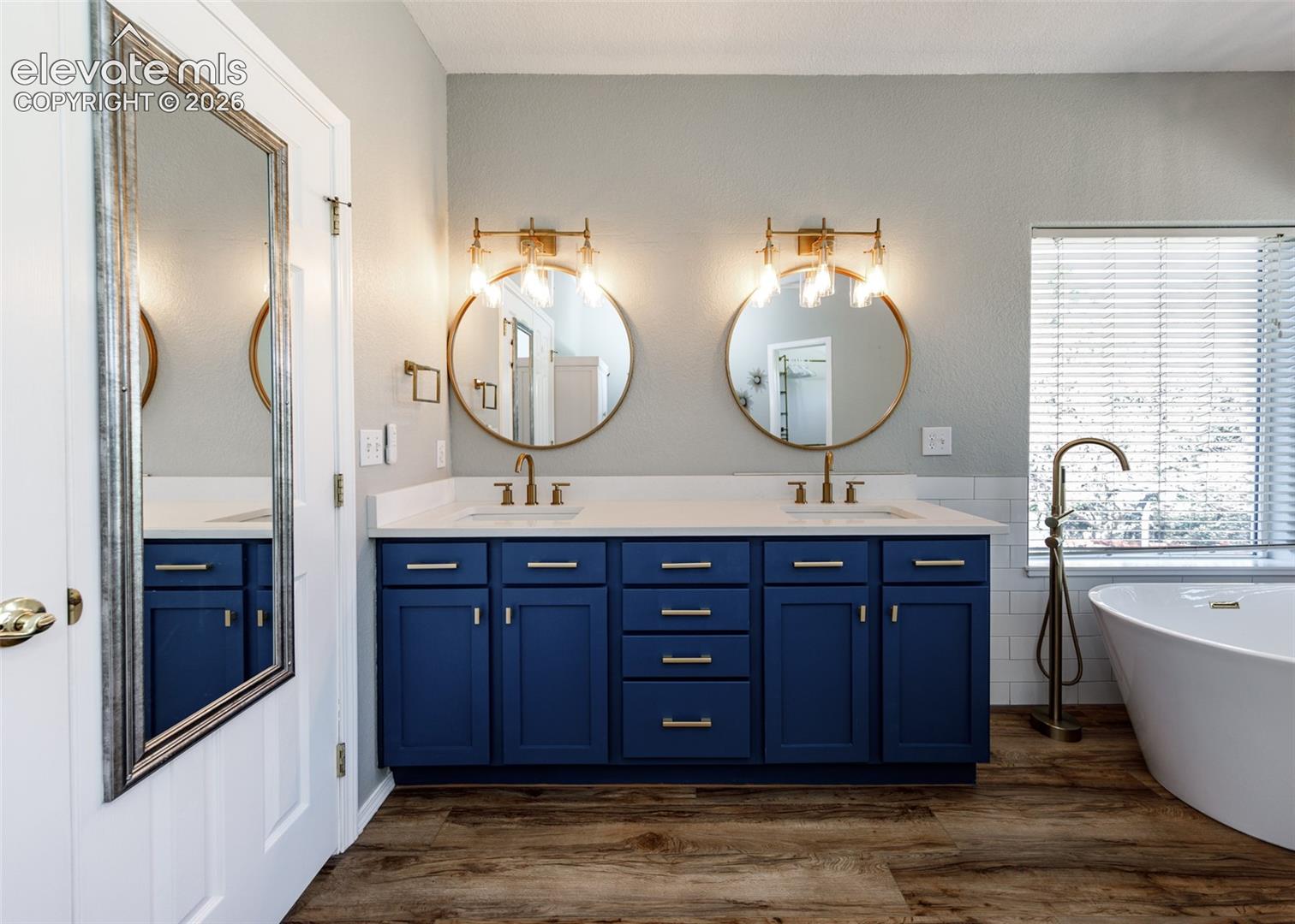 Double vanity featuring deep blue cabinetry, quartz countertops, dual circular gold-framed mirrors, wall-mounted light fixtures, and a standalone soaking tub with a floor-mounted gold faucet