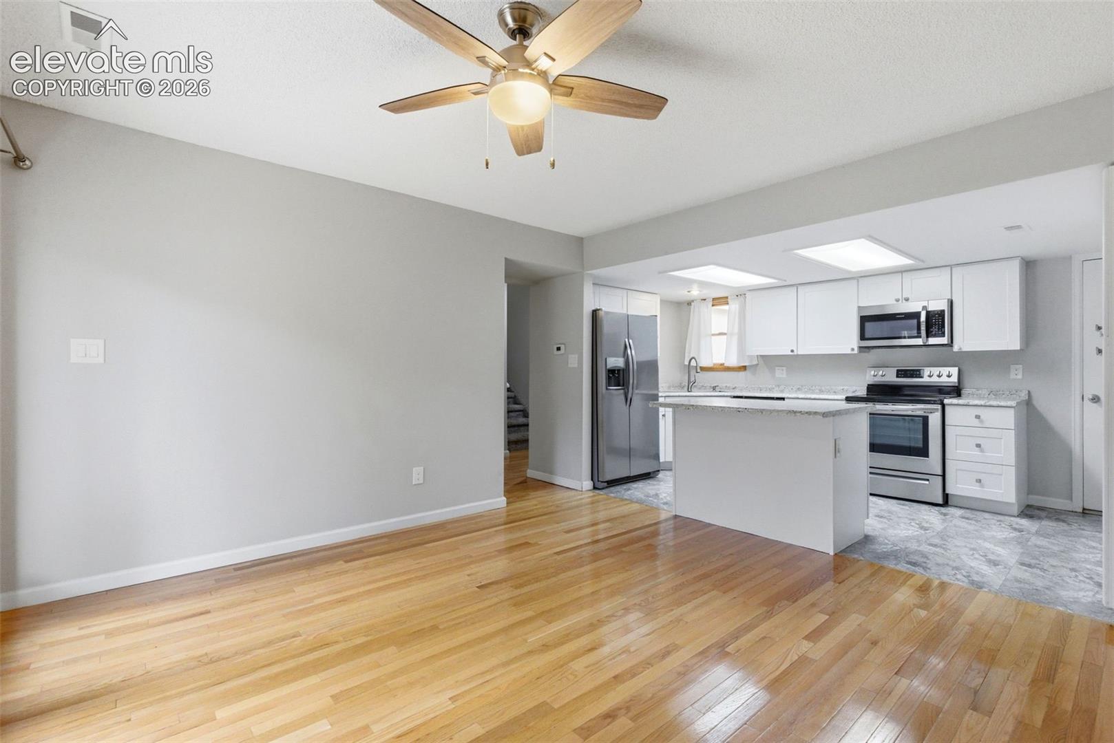 Open-concept living space featuring wood-finish flooring, a ceiling fan with integrated lighting, and a kitchen area with white cabinetry, stainless steel appliances, and a central island