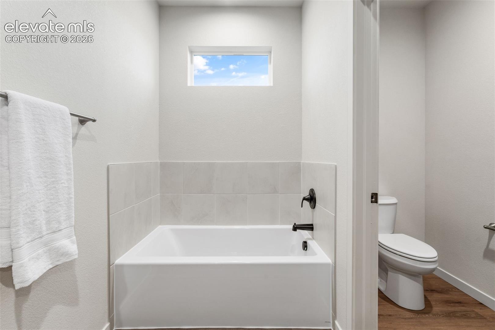 Bathroom featuring a deep soaking tub with a tiled surround, transom window, matte black plumbing fixtures, and wood-finish flooring