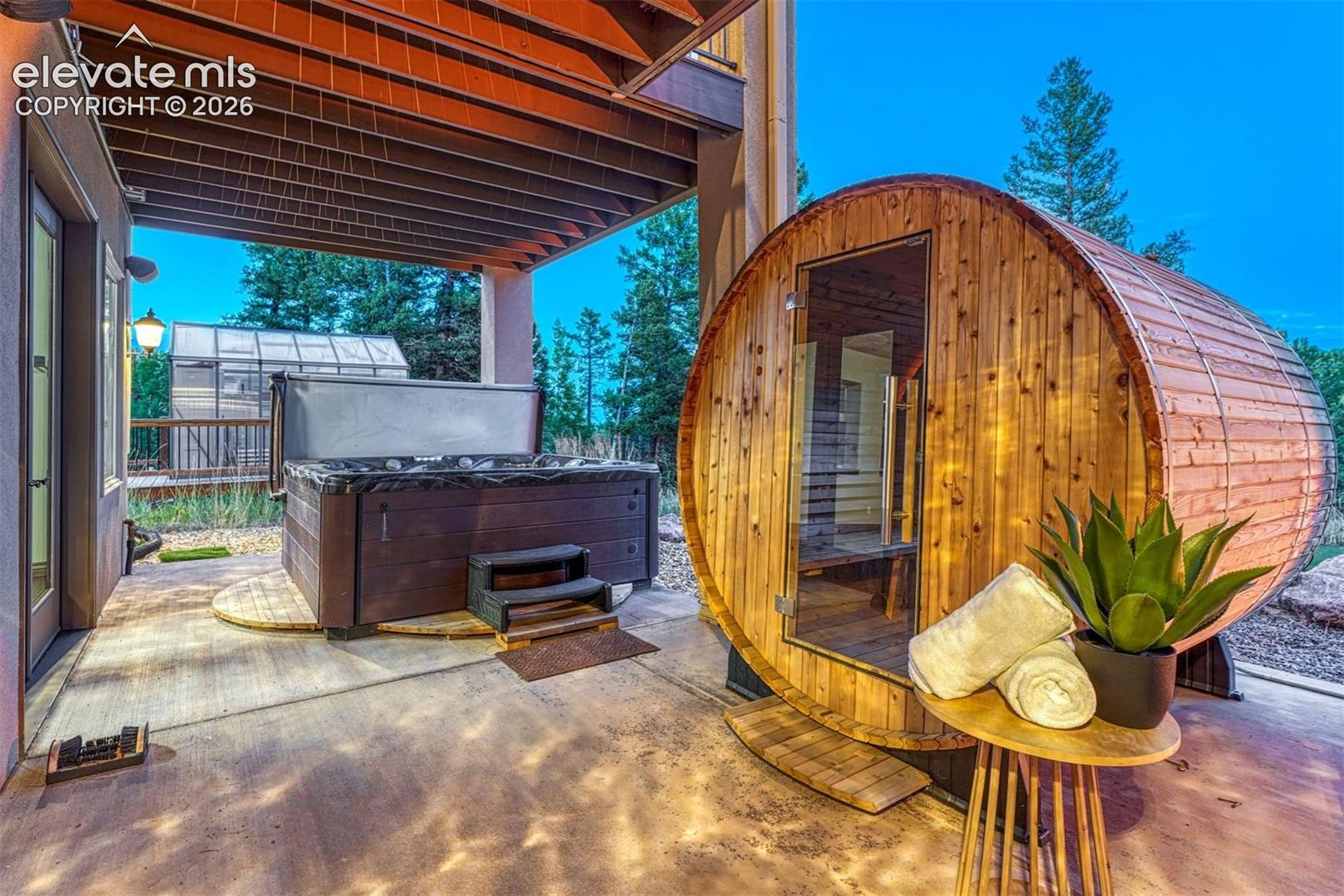 Expansive outdoor patio with a built-in hot tub, barrel-style sauna, and a prominent greenhouse structure