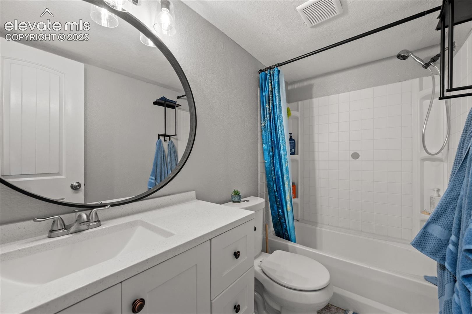 Bathroom vanity featuring a solid-surface countertop with an integrated rectangular sink, large circular wall-mounted mirror, matte black hardware, and a tub-shower combination with built-in shelving