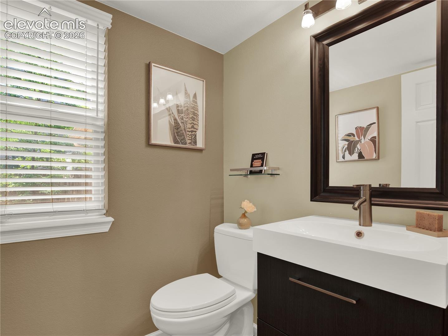 Wall-mounted vanity with a rectangular sink, dark wood-finish cabinetry, metal hardware, framed mirror with integrated light fixture, and window with shutter blinds