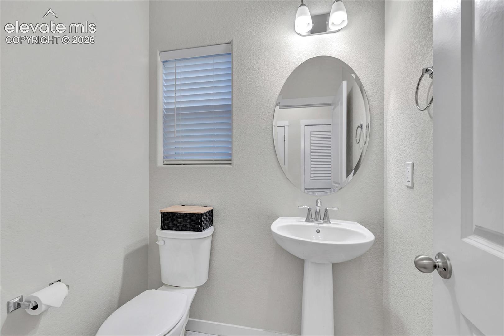 Pedestal sink with chrome faucet, oval mirror, wall-mounted light fixture, and window with blinds