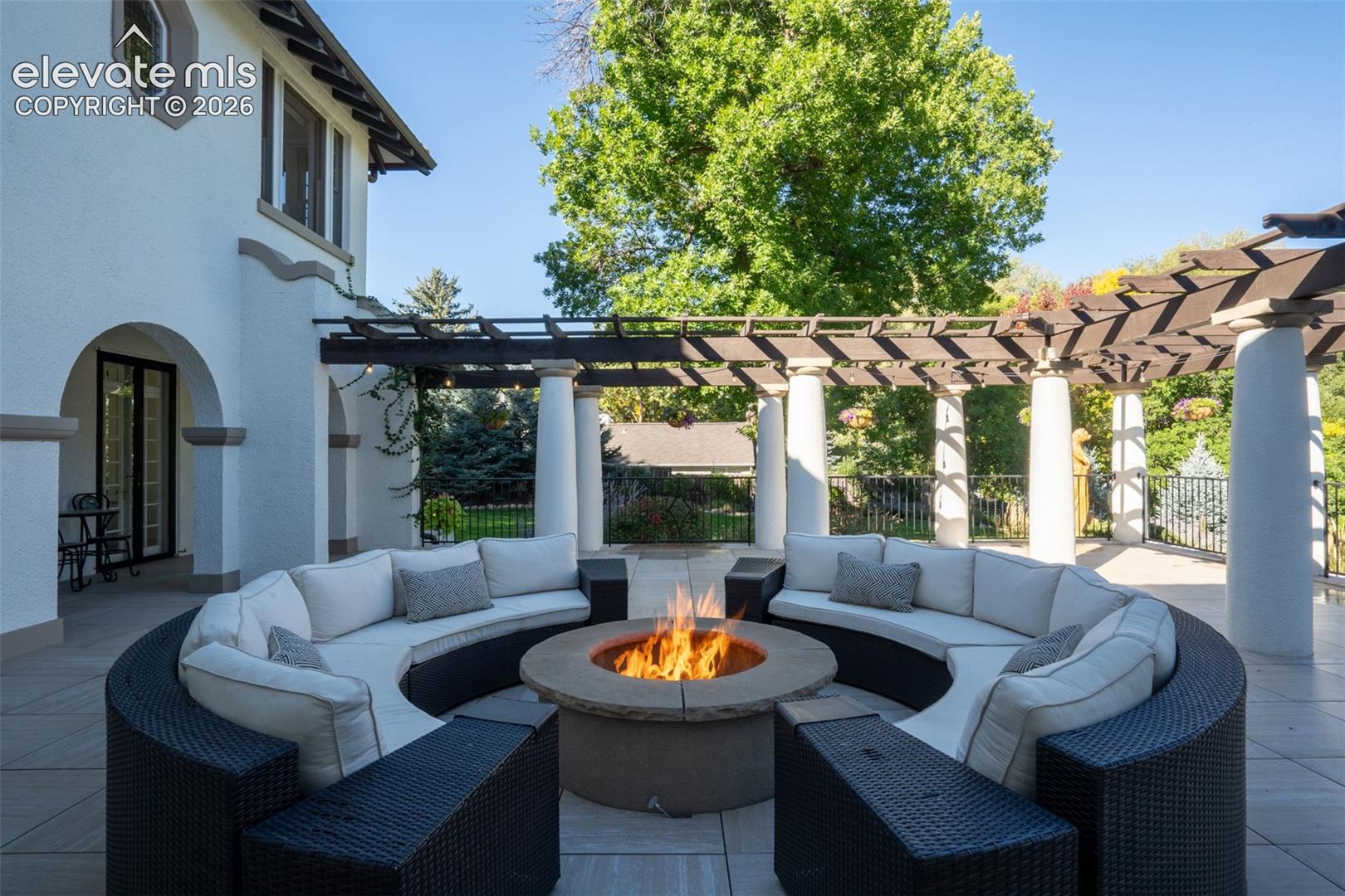 Expansive stone-paved patio featuring a circular permanent fire pit, a curved pergola, and structural columns