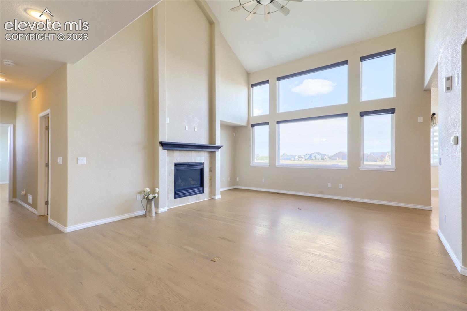 Dramatic two-story great room featuring soaring vaulted ceilings, a modern ceiling fan, and a floor-to-ceiling accent wall with a gas fireplace, tile surround, and mantel. Expansive two-tier wall-of-windows floods the open floor plan with natural light while capturing expansive outdoor views, complemented by hardwood flooring.