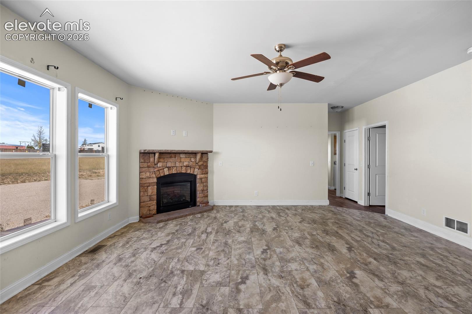 Living area featuring stone-surround fireplace with mantel, wood-finish flooring, ceiling fan with integrated lighting, and large windows