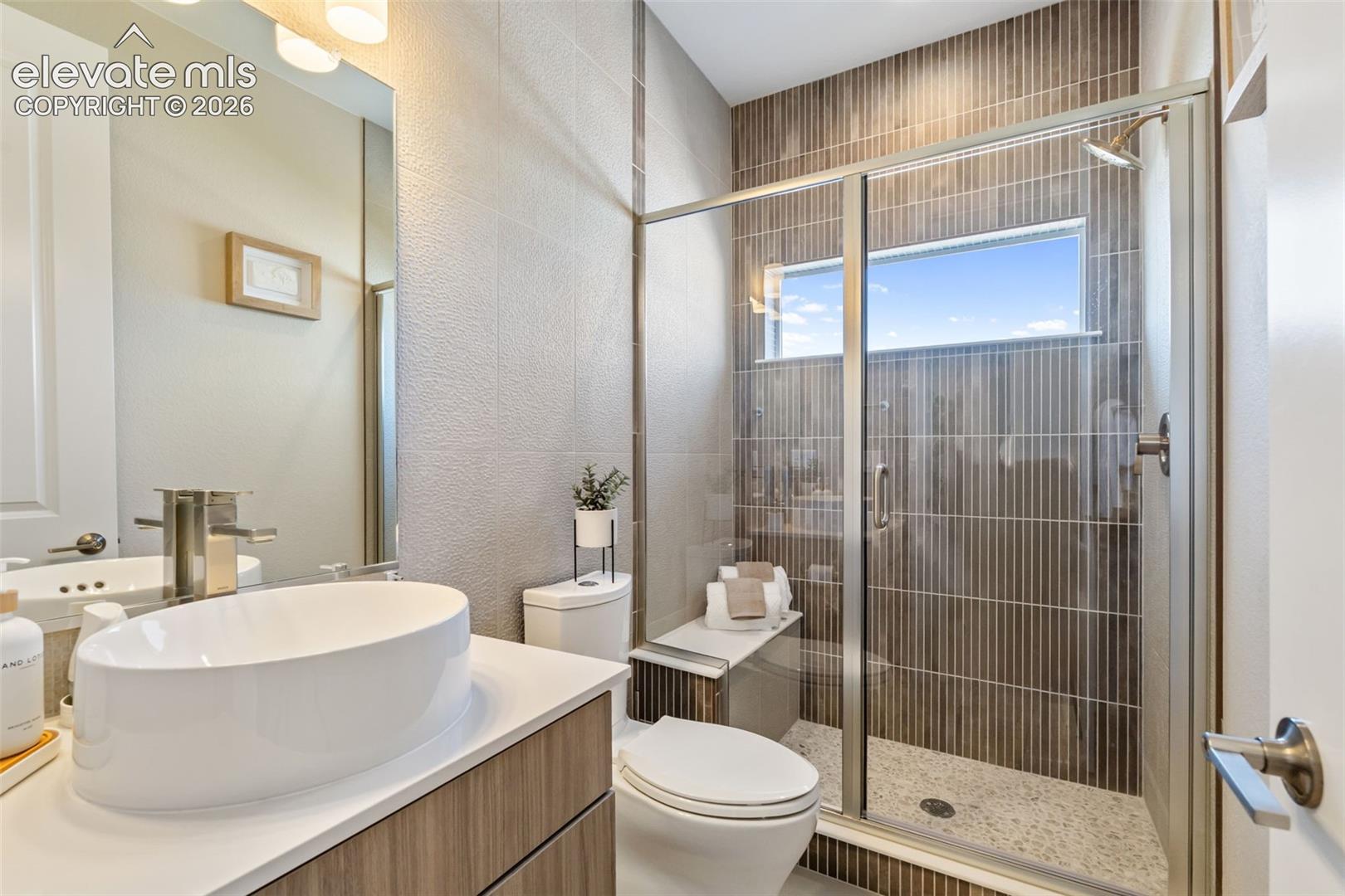 Bathroom featuring a wood-finish vanity with a white vessel sink, quartz countertop, glass-enclosed shower with vertical wall tiling, and a built-in shower bench