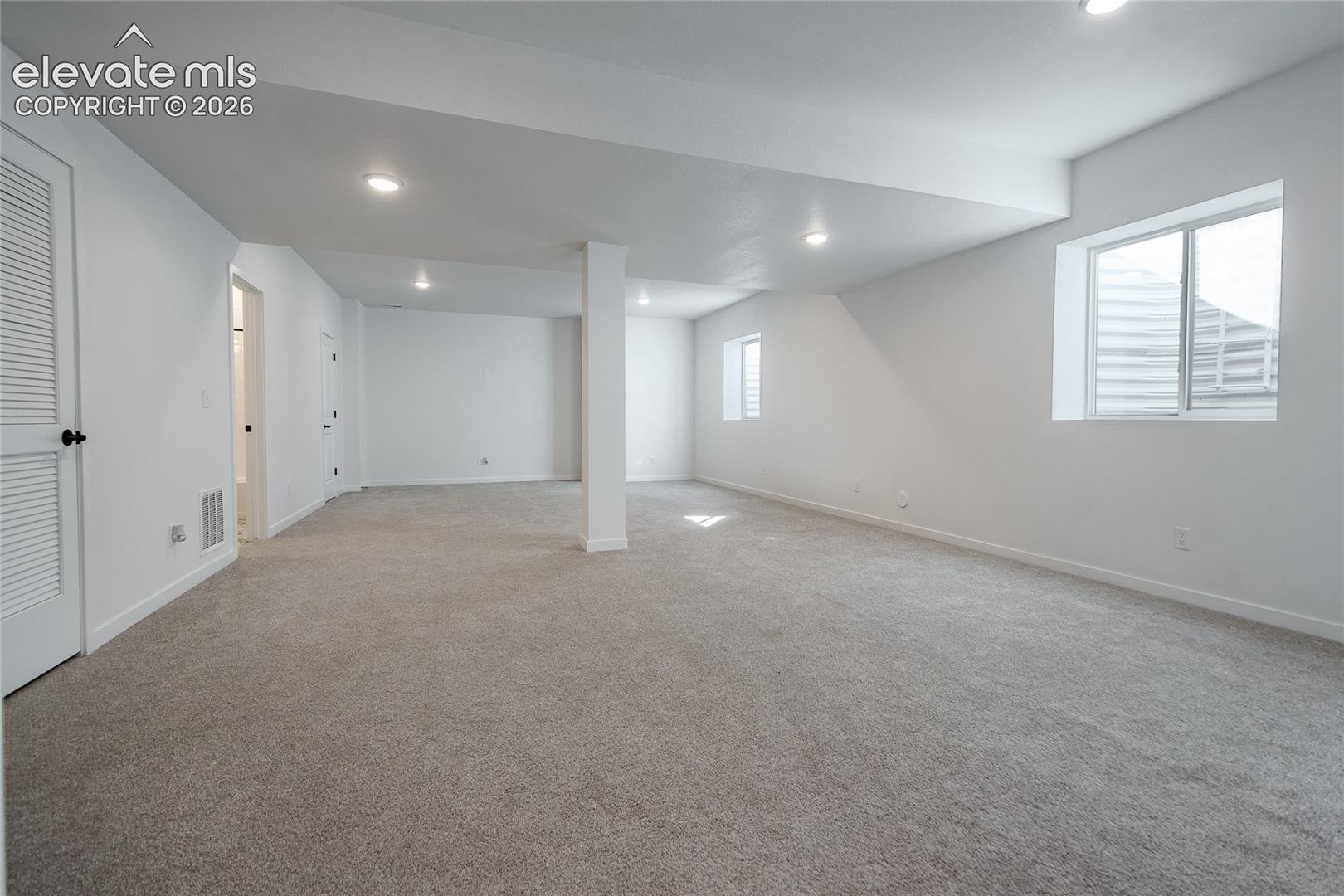 Open-concept interior featuring neutral wall-to-wall carpeting, recessed ceiling lighting, louvered closet doors, and multiple windows providing natural light